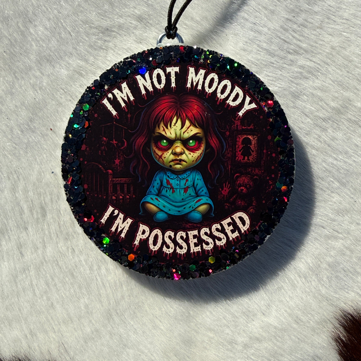 A round black glitter car freshie featuring a creepy illustrated girl with red hair and the text 'I'M NOT MOODY I'M POSSESSED' in white, hanging on a black string against a white textured background.
