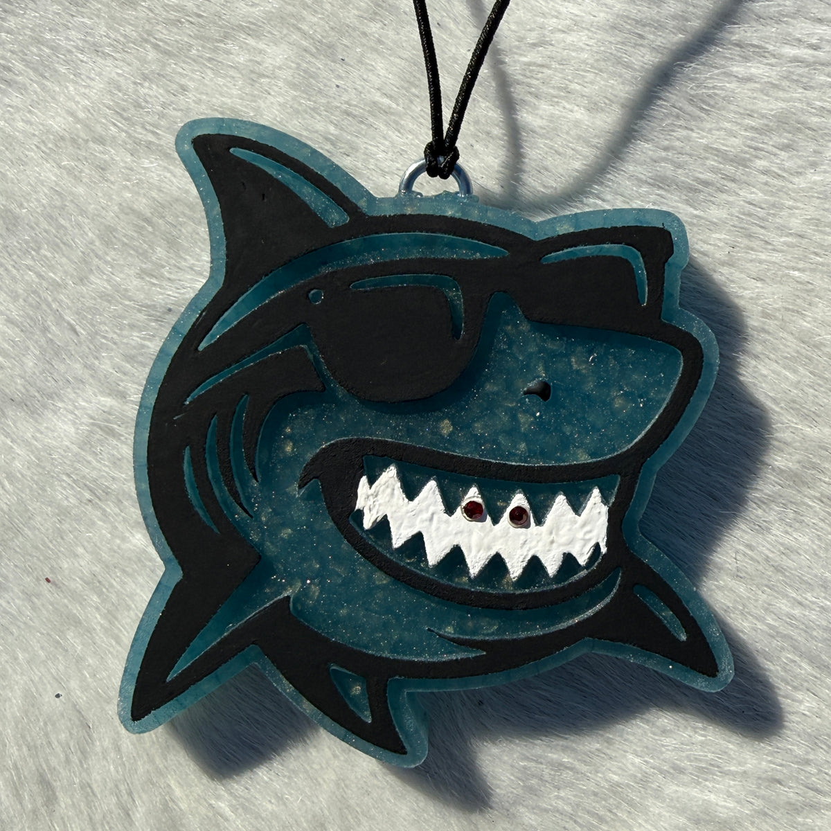 A shark-shaped car freshie with a black outline and a teal-blue center, featuring a black sunglasses graphic, white teeth, and a black hanging cord on a light textured background.