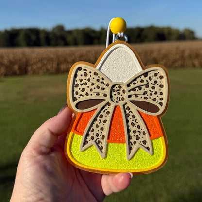 A hand holds a candy corn-shaped car freshie outdoors against a blurred field and sky background. The car freshie has a beige bow with dark spots, orange and yellow glitter sections, and a silver hanging loop at the top.