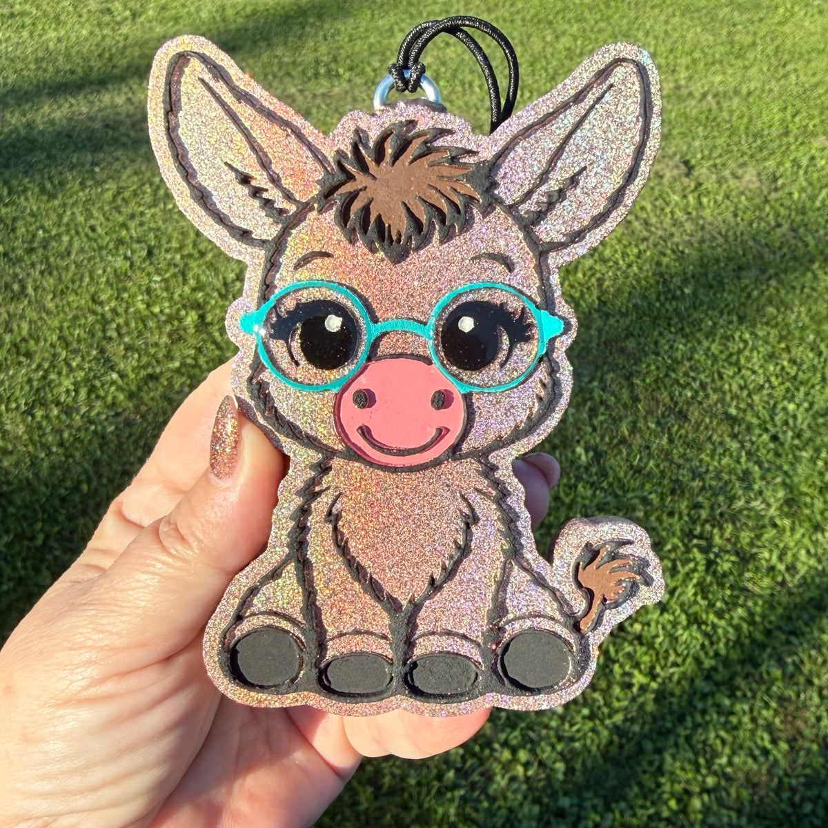 A hand holds a glittery donkey-shaped car freshie outdoors on a green grass background. The donkey has turquoise glasses, a pink snout, and black hooves.