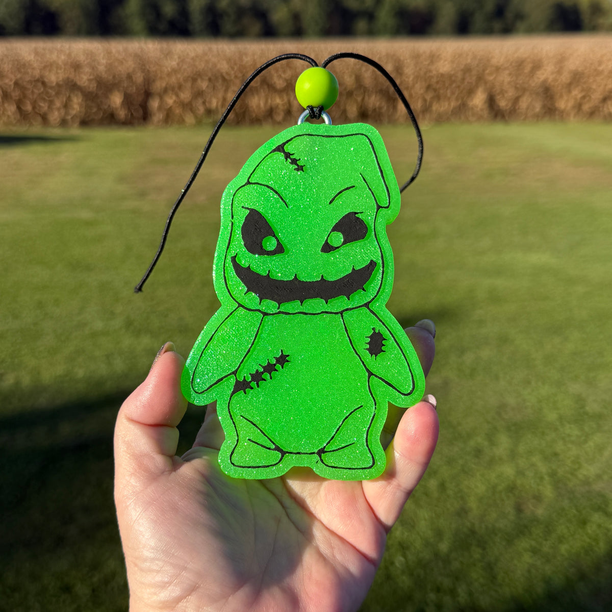 A hand holds a bright green glittery character-shaped air freshener or car freshie with black stitched details and a black hanging cord, set against an outdoor grassy field background.