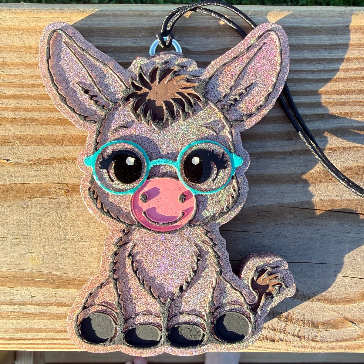 A glittery cartoon donkey-shaped car freshie with turquoise glasses and a pink snout, shown on a wooden surface with a black cord attached.