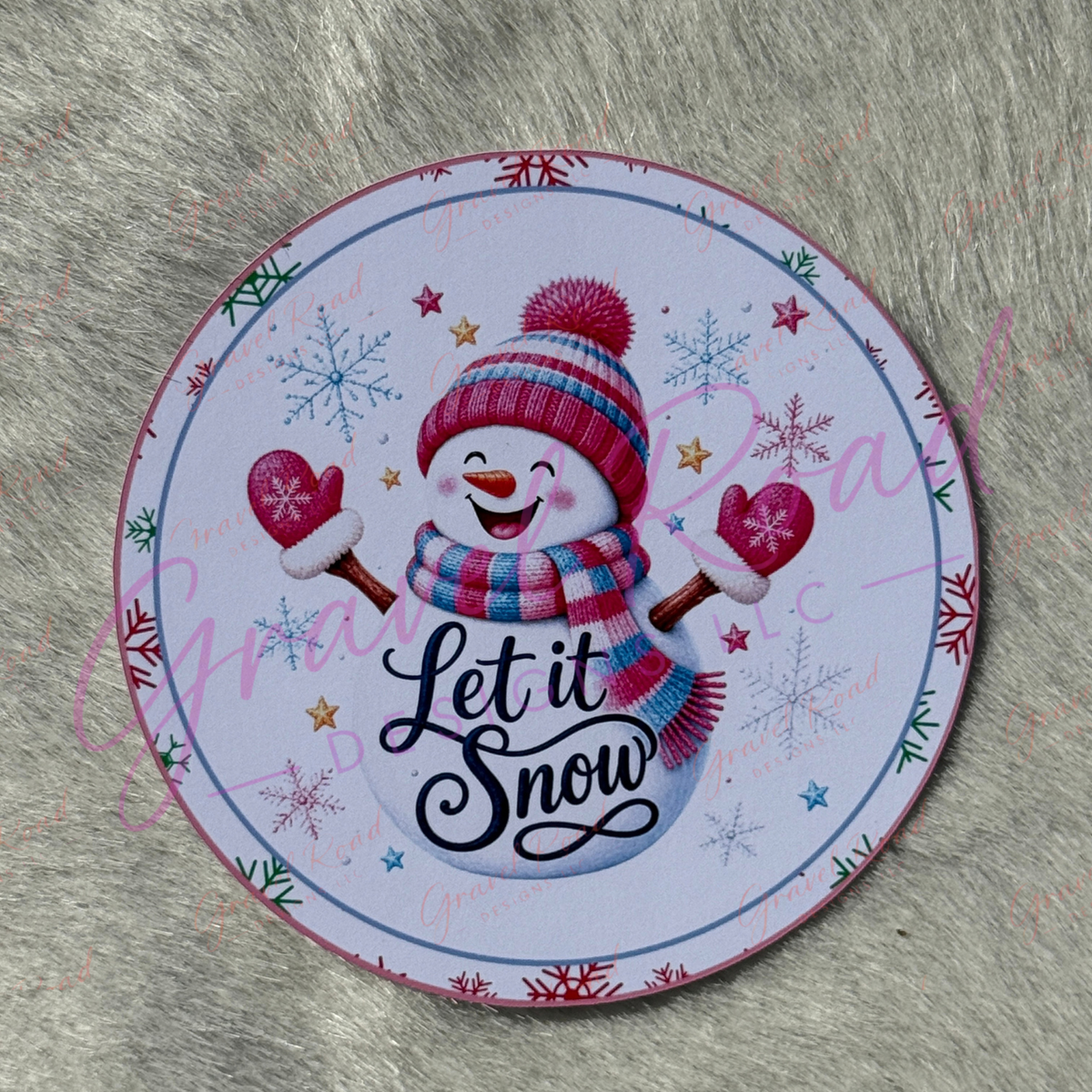 A round printed cardstock featuring a festive snowman illustration with the words “Let it Snow,” placed on a gray faux-fur surface. The design includes a pink, blue, and white snowman, snowflakes, stars, and a blue border. Displayed on a faux fur background
