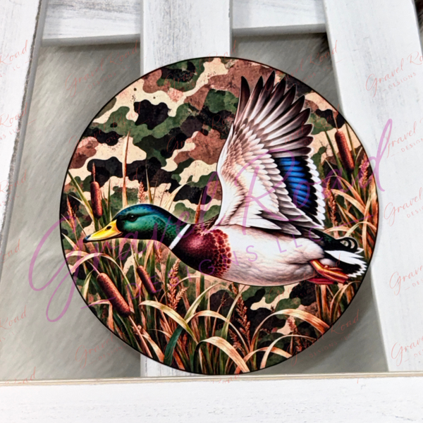 A round cardstock featuring a detailed illustration of a mallard duck flying over marsh grasses with a camouflage background, displayed on a white wooden surface.