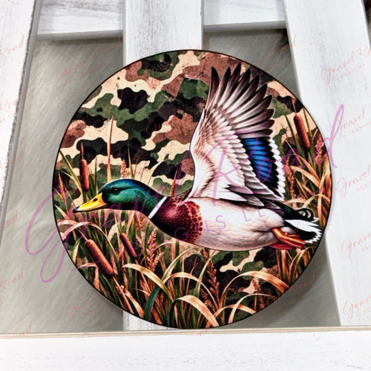 A round cardstock featuring a detailed illustration of a mallard duck flying over marsh grasses with a camouflage background, displayed on a white wooden surface.