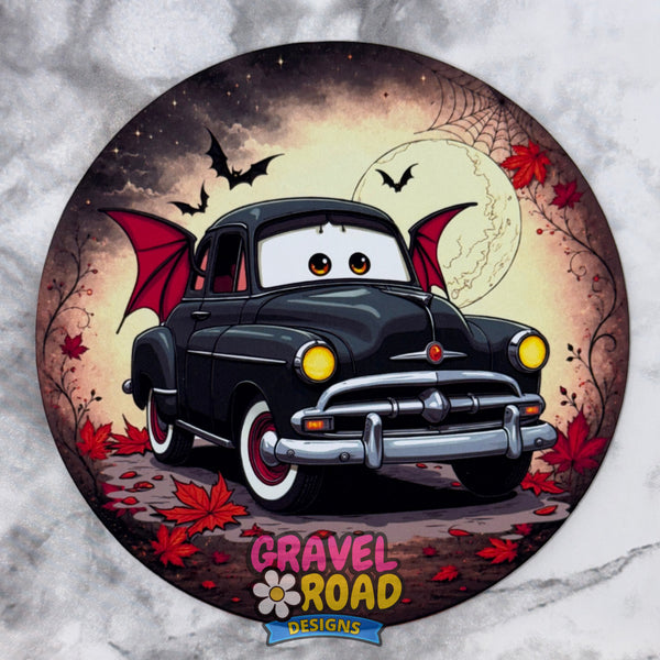 A circular cardstock featuring a cartoon-style black vintage car with red wings and eyes, set against a spooky full moon and bats background. The design includes red leaves and the logo 'GRAVEL ROAD DESIGNS' at the bottom, shown on a light marble-like surface.