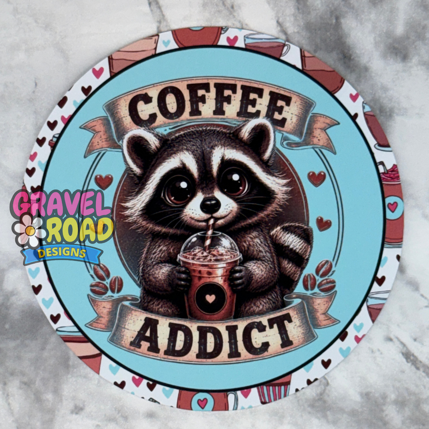 A round printed cardstock featuring a cute raccoon holding a coffee drink with the words 'COFFEE ADDICT' on a light blue background, surrounded by a patterned border. A 'Gravel Road Designs' logo is visible on the left side.