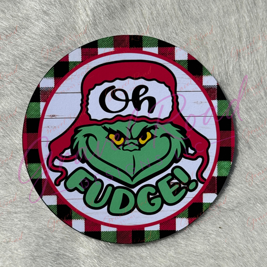 A circular cardstock featuring the green face wearing a red Santa hat with 'Oh Fudge!' text, set on a black, white, and green plaid background with a faux fur backdrop.