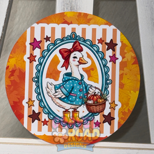 Round fall cardstock featuring a white goose in a teal polka-dot coat and red bow, holding a basket of pumpkins.