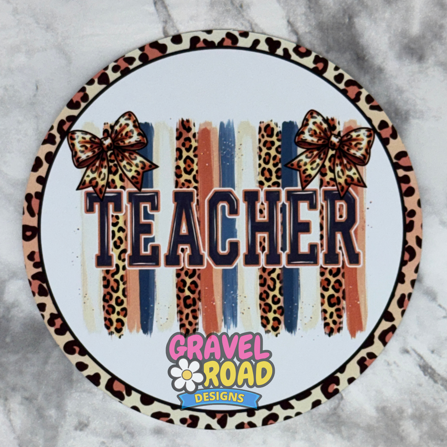 A round cardstock featuring the word “TEACHER” in large outlined letters over vertical leopard-print and striped brushstroke bands, with two leopard bows and a border. The bottom includes the logo “GRAVEL ROAD DESIGNS.
