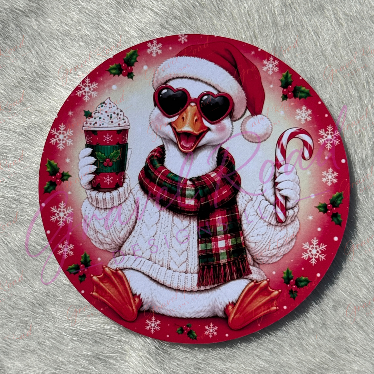 A round Christmas-themed cardstock featuring a white duck wearing a red Santa hat, heart-shaped sunglasses, a red and green plaid scarf, and a white knit sweater. The duck holds a hot cocoa drink and a candy cane, surrounded by red and pink holiday patterns with snowflakes, holly, and white snowflakes. Displayed on a faux fur background.