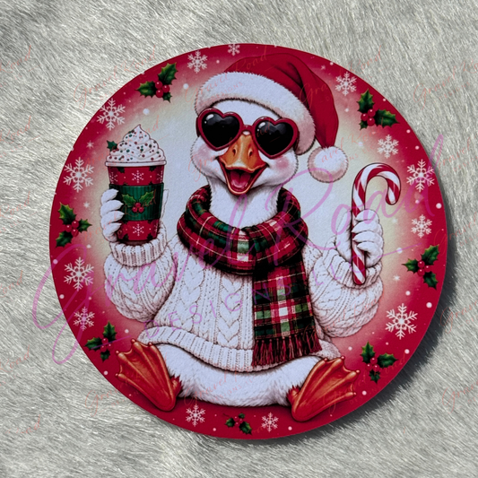 A round Christmas-themed cardstock featuring a white duck wearing a red Santa hat, heart-shaped sunglasses, a red and green plaid scarf, and a white knit sweater. The duck holds a hot cocoa drink and a candy cane, surrounded by red and pink holiday patterns with snowflakes, holly, and white snowflakes. Displayed on a faux fur background.