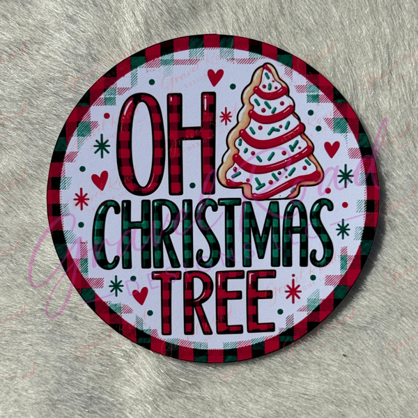 A round cardstock featuring the phrase 'OH CHRISTMAS TREE' in red and green lettering, with a decorated Christmas tree illustration and a red-and-green plaid border, placed on a gray faux-fur surface.