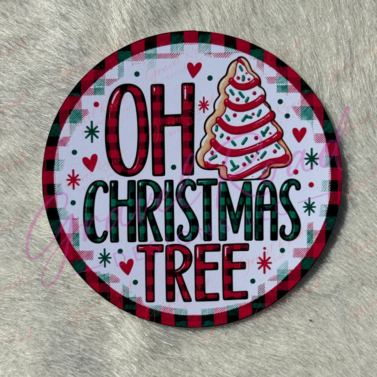 A round cardstock featuring the phrase 'OH CHRISTMAS TREE' in red and green lettering, with a decorated Christmas tree illustration and a red-and-green plaid border, placed on a gray faux-fur surface.