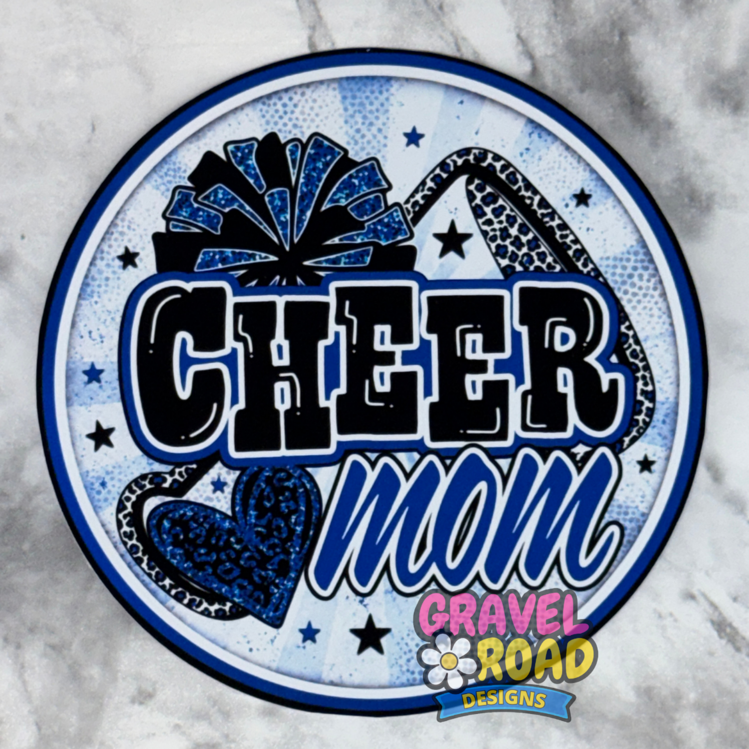 A circular cardstock design featuring the words 'CHEER mom' in bold black and blue lettering, decorated with a blue leopard-print pom-pom, a blue leopard-print heart, and small stars on a light blue patterned background. The design includes a 'Gravel Road Designs' logo at the bottom.