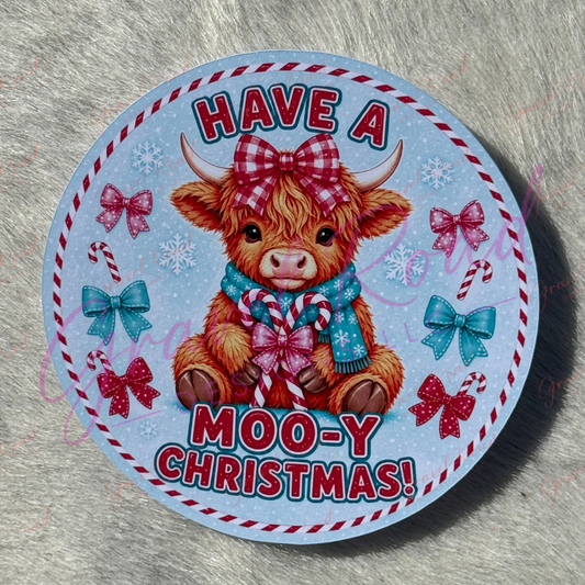 A round cardstock featuring a cute brown Scottish highland cow with a red plaid bow and blue scarf, surrounded by candy canes, snowflakes, bows, and the text 'HAVE A MOO-Y CHRISTMAS!' on a light blue background, placed on a gray faux fur surface.