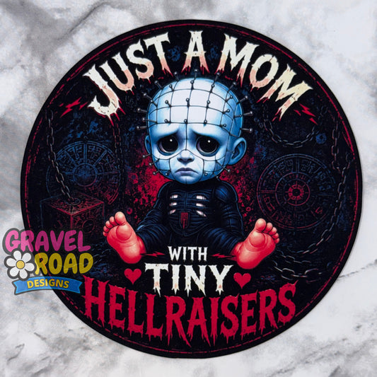 A circular cardstock design featuring a stylized blue horror character with red feet and the text 'JUST A MOM WITH TINY HELLRAISERS' on a dark background, placed on a light marble-like surface. A 'Gravel Road Designs' logo appears in the lower left.