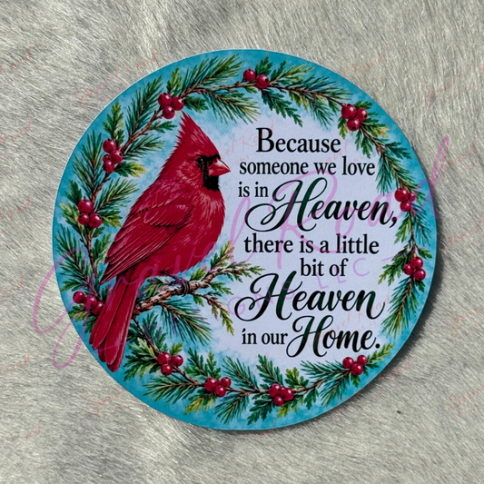 A round cardstock featuring a red cardinal bird, green pine branches, red berries, and black script text "Because someone we love is in Heaven, there is a little bit of Heaven in our home" on a light blue background, photographed on a gray textured surface.