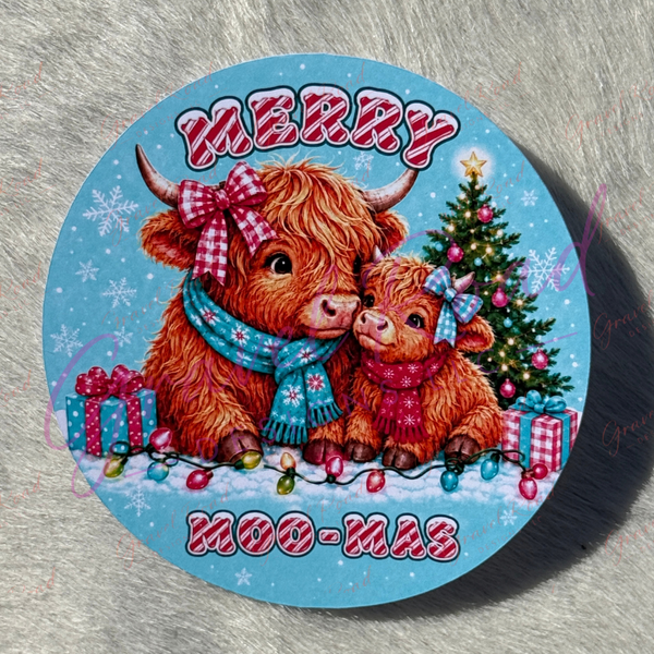 A round printed cardstock featuring a festive Christmas illustration with two Highland cows, a decorated tree, gifts, and the words “MERRY MOO-MAS” on a light blue background, photographed on a gray textured surface.