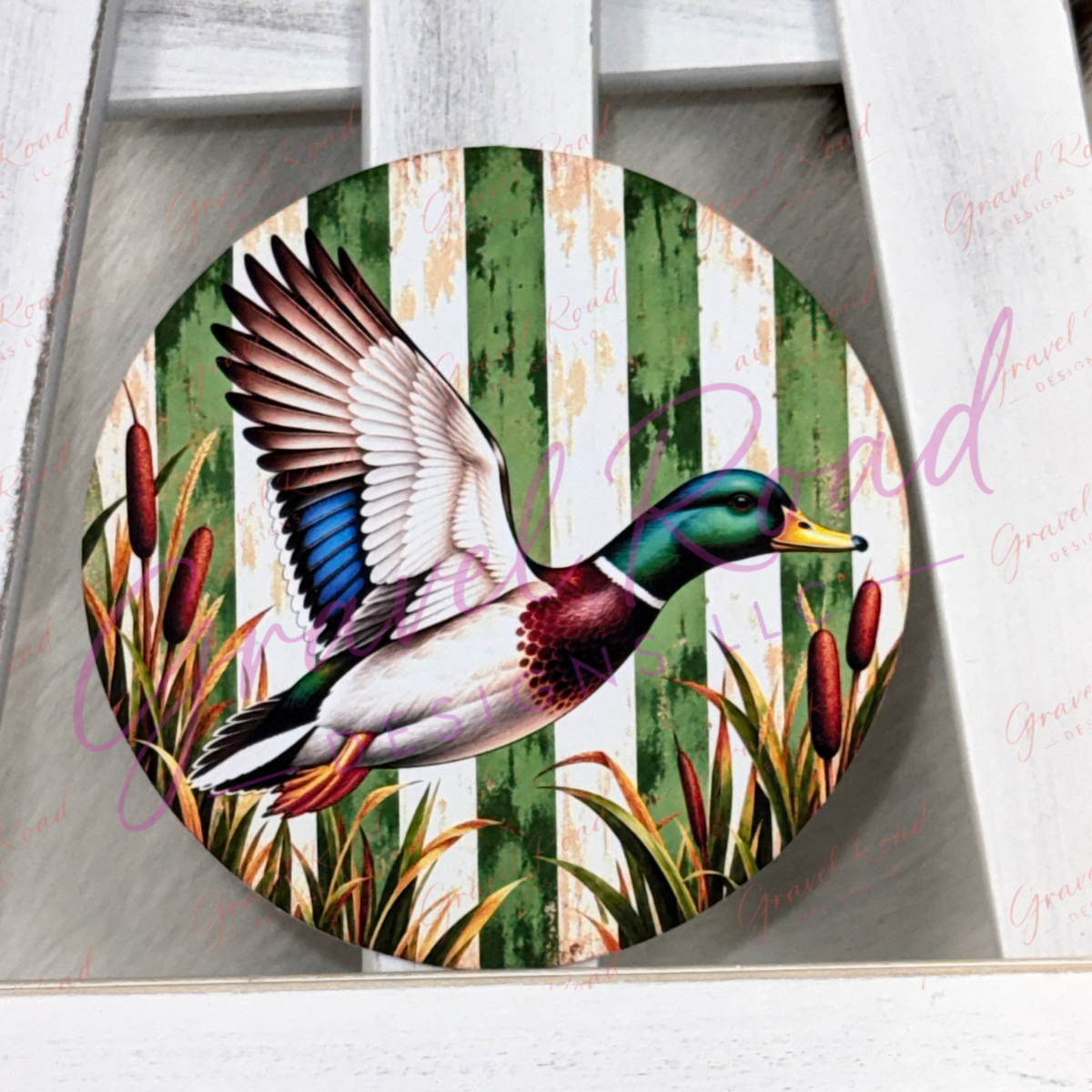 Round cardstock featuring a flying mallard duck over green and white vertical stripes with cattails, displayed on a white wooden surface.