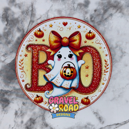 A circular Halloween-themed cardstock featuring the word 'BOO' in glittery red letters with a cute white ghost holding a jack-o'-lantern, plus small pumpkins and stars. The bottom reads 'Gravel Road Designs' on a blue banner. On a grey marble background.