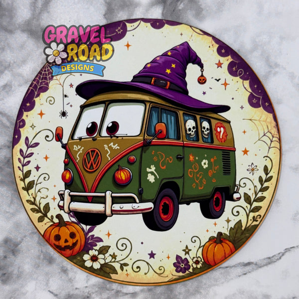 A circular cardstock featuring a cartoon VW bus decorated for Halloween with a purple witch hat, skulls, pumpkins, flowers, and spiderweb motifs. The top left has the logo 'GRAVEL ROAD DESIGNS' and the bus has a visible Volkswagen logo. On a grey marble background.