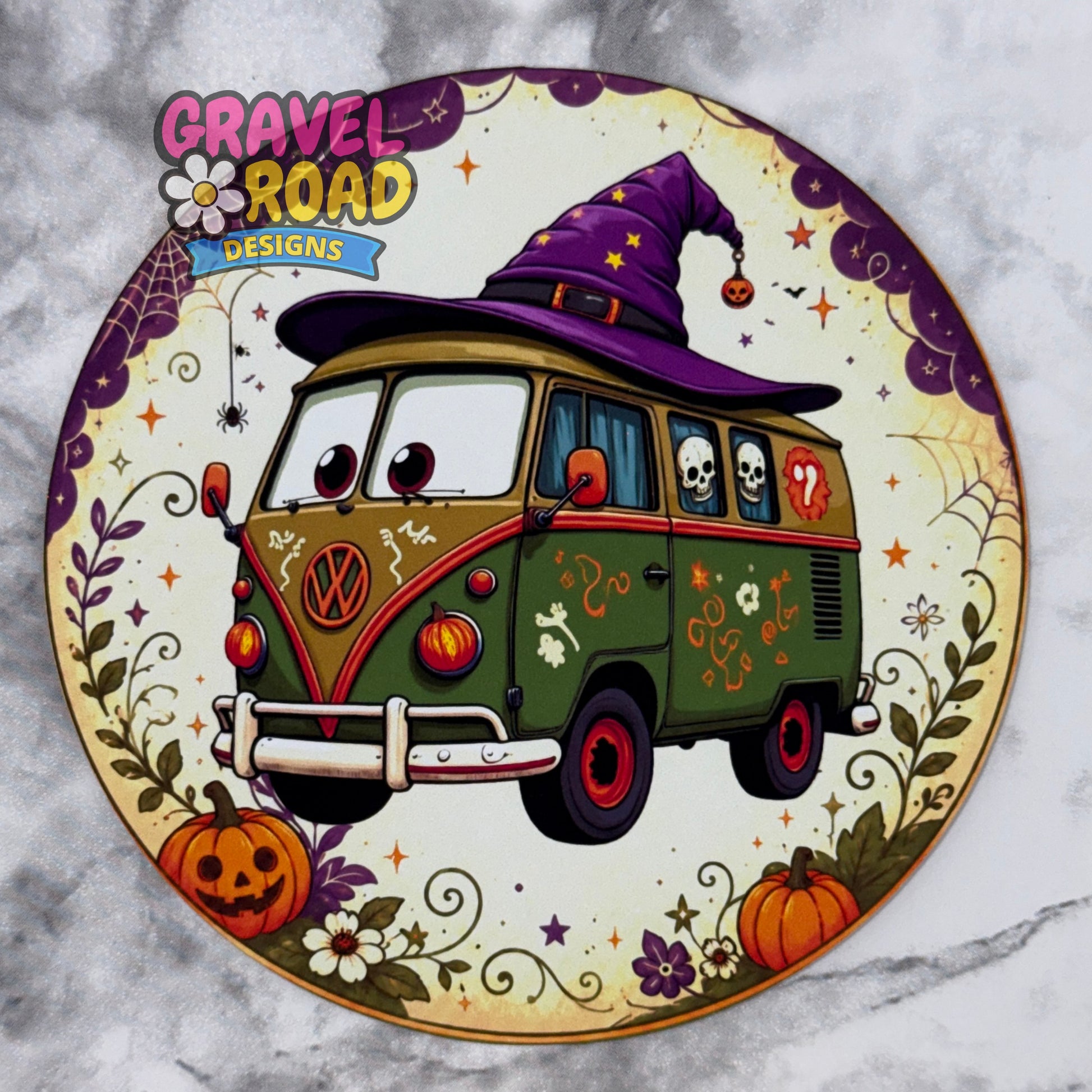 A circular cardstock featuring a cartoon VW bus decorated for Halloween with a purple witch hat, skulls, pumpkins, flowers, and spiderweb motifs. The top left has the logo 'GRAVEL ROAD DESIGNS' and the bus has a visible Volkswagen logo. On a grey marble background.