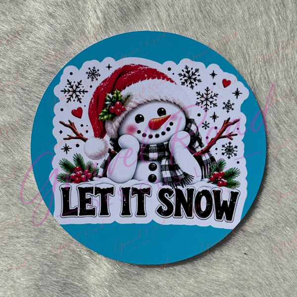 A round cardstock featuring a cartoon snowman wearing a red Santa hat and plaid scarf, surrounded by snowflakes, berries, and the words 'LET IT SNOW' on a bright blue background, placed on a gray textured surface.