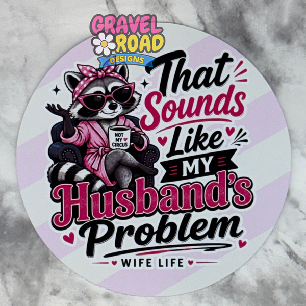 A round cardstock featuring a cartoon raccoon in a pink robe and sunglasses holding a mug that says 'NOT MY W CIRCUS,' with the text 'That Sounds Like My Husband's Problem' and 'Wife Life' below. The design includes the brand/logo 'Gravel Road Designs' at the top.