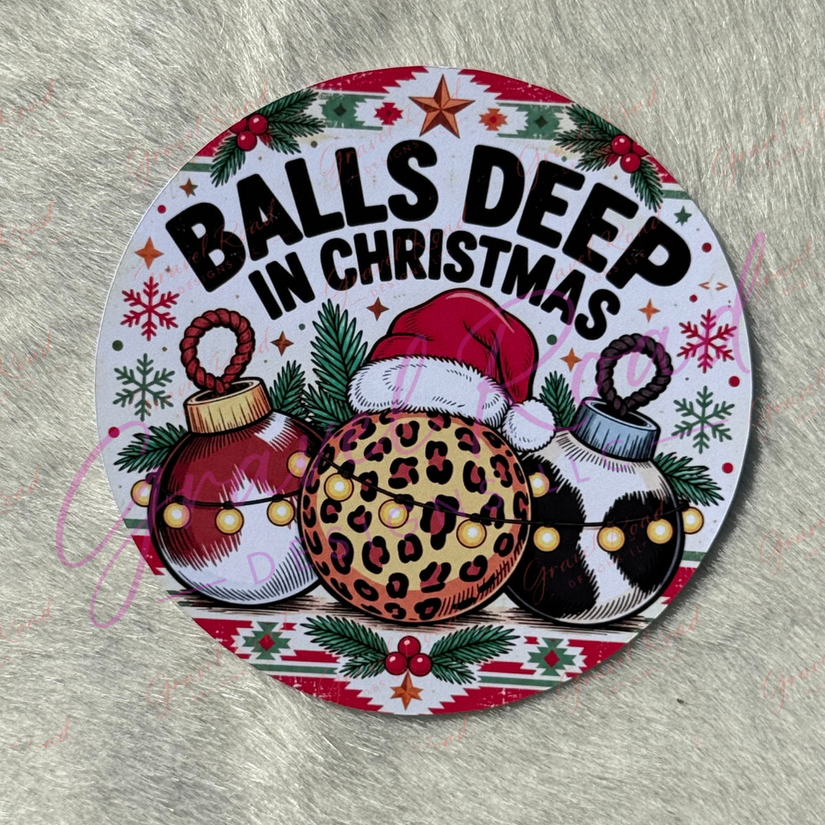 A round printed cardstock featuring the phrase “BALLS DEEP IN CHRISTMAS” above three Christmas ornaments. The design includes pine branches, snowflakes, red berries, a red Santa hat, and a leopard-print ornament in the center, and two cow prints ornaments. Placed on a light gray furry surface.