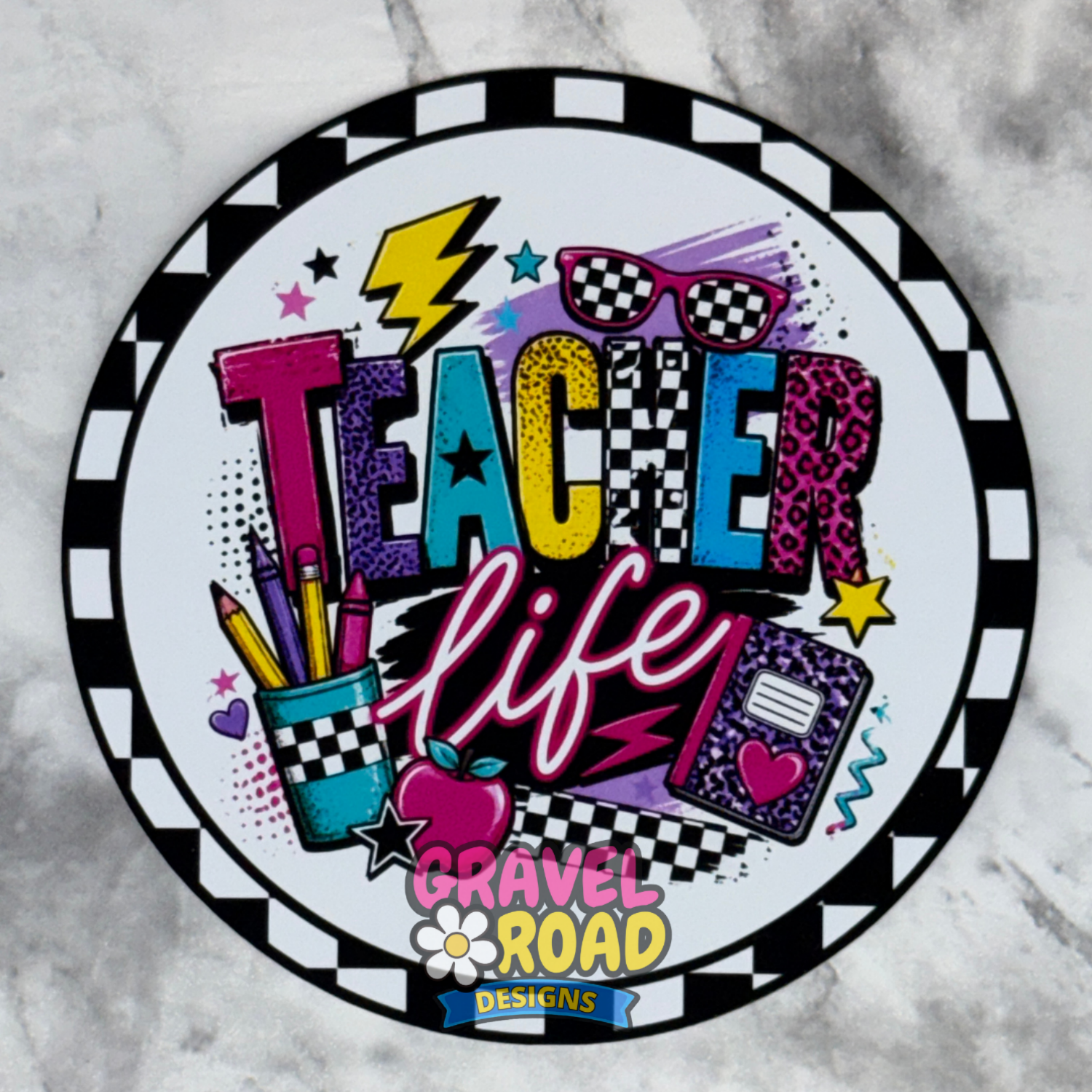 A circular cardstock design featuring the words 'Teacher Life' in colorful retro-style lettering, surrounded by school-themed doodles like pencils, an apple, sunglasses, and a notebook. The design includes a black-and-white checkered border and a 'Gravel Road Designs' logo at the bottom, shown on a light gray marble surface.