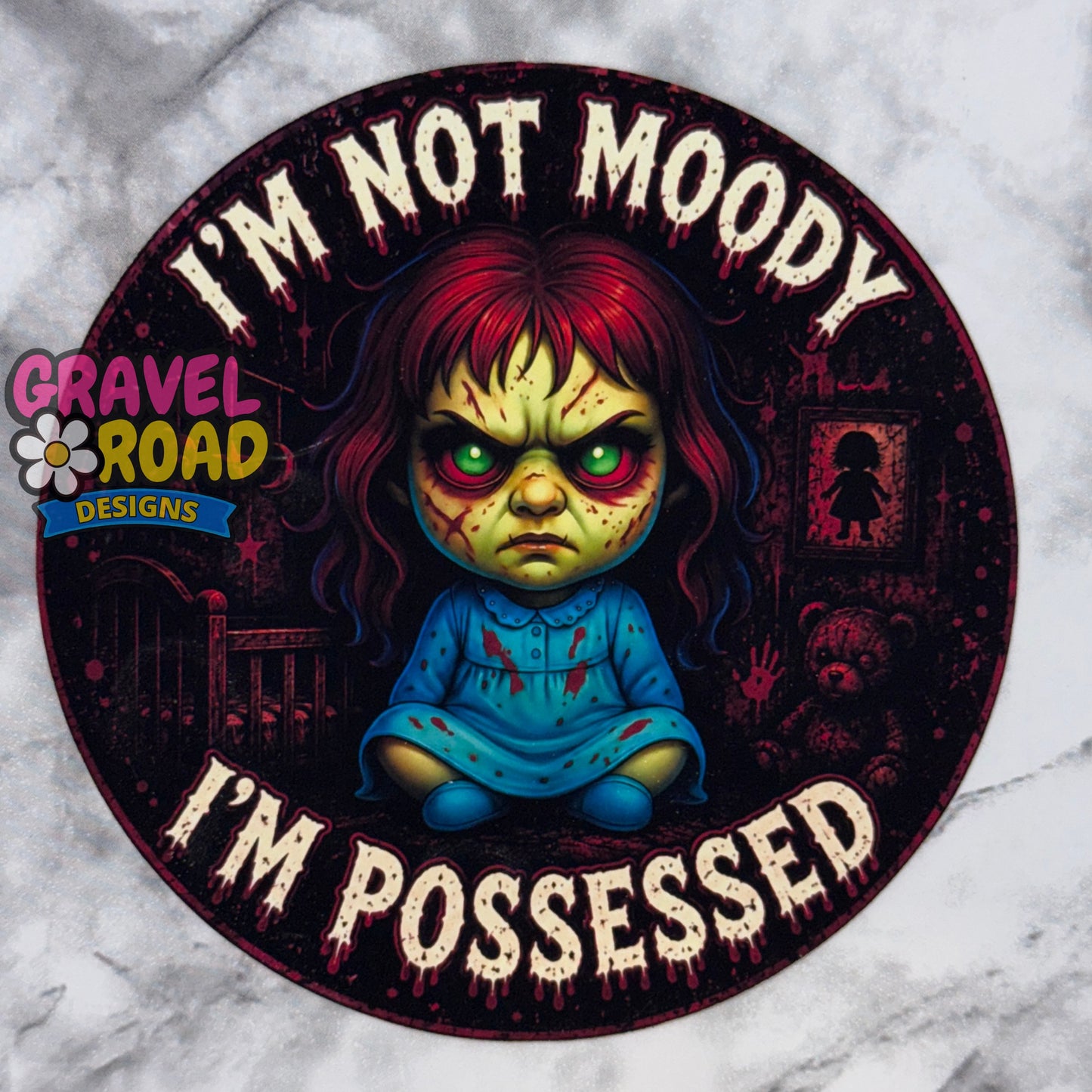 A circular cardstock featuring a creepy cartoon girl with red hair, green eyes, and a blue dress, surrounded by the text 'I'M NOT MOODY I'M POSSESSED' in dripping white letters. A 'Gravel Road Designs' logo is visible on the left side.