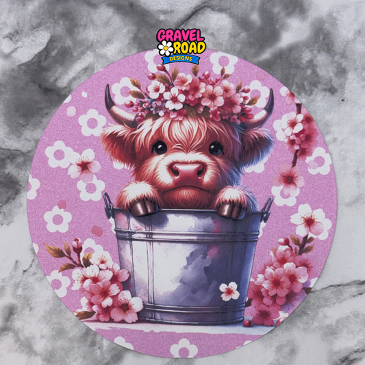 Circular design with a cartoon cow in a bucket surrounded by flowers on a pink background, branded 'Gravel Road'.