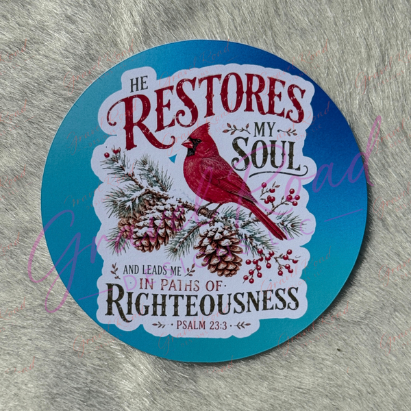 A round cardstock featuring a red cardinal perched on pine branches with pinecones and berries, set against a blue background with the text 'HE RESTORES MY SOUL AND LEADS ME IN PATHS OF RIGHTEOUSNESS' and 'PSALM 23:3'. The cardstock is photographed on a textured gray surface.