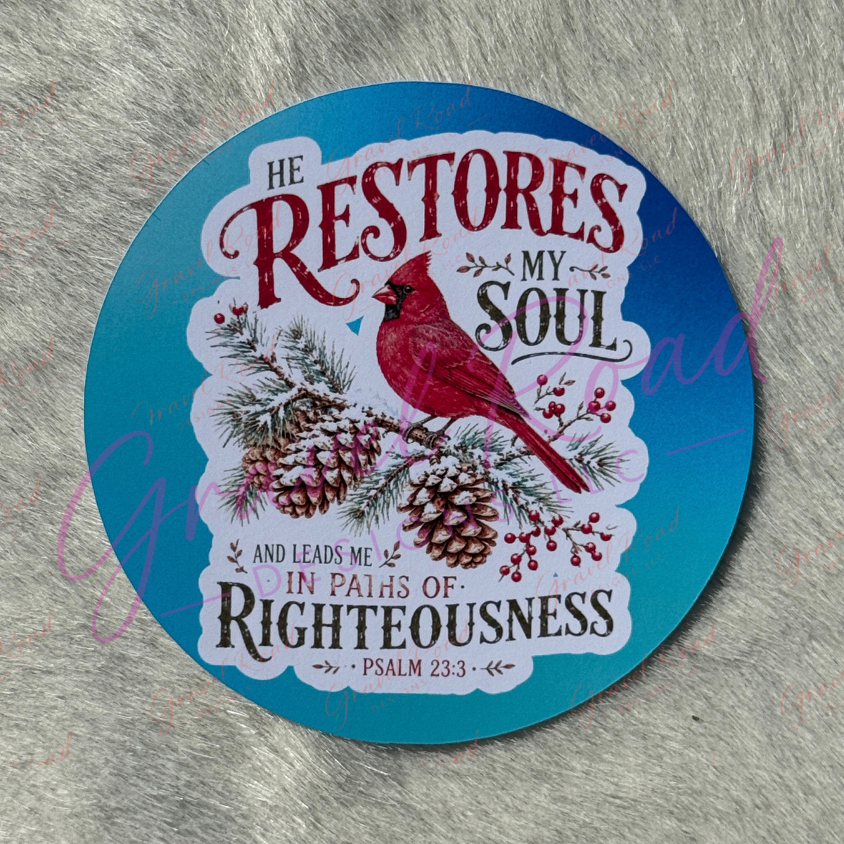 A round cardstock featuring a red cardinal perched on pine branches with pinecones and berries, set against a blue background with the text 'HE RESTORES MY SOUL AND LEADS ME IN PATHS OF RIGHTEOUSNESS' and 'PSALM 23:3'. The cardstock is photographed on a textured gray surface.