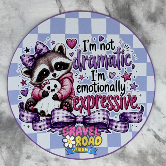 A circular cardstock design featuring a cute cartoon raccoon holding a small ghost, with the text 'I'm not dramatic, I'm emotionally expressive.' and a purple plaid ribbon bow.