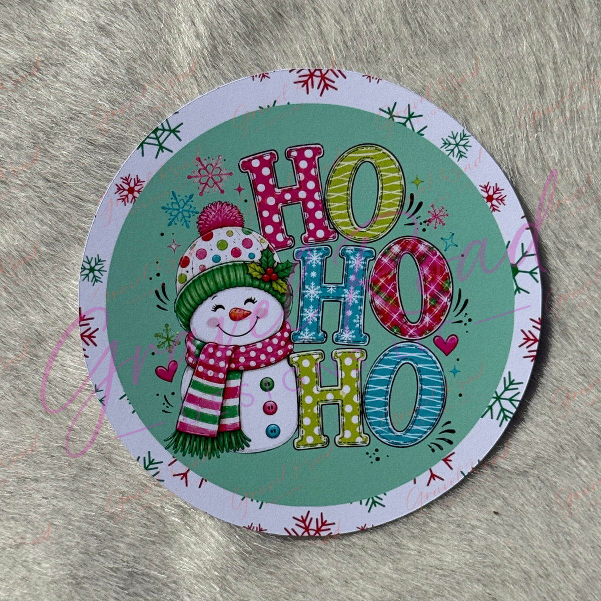 A round printed cardstock featuring a festive 'HO HO HO' holiday design with a cartoon snowman wearing a green hat and red polka-dot scarf, placed on a gray faux-fur surface.