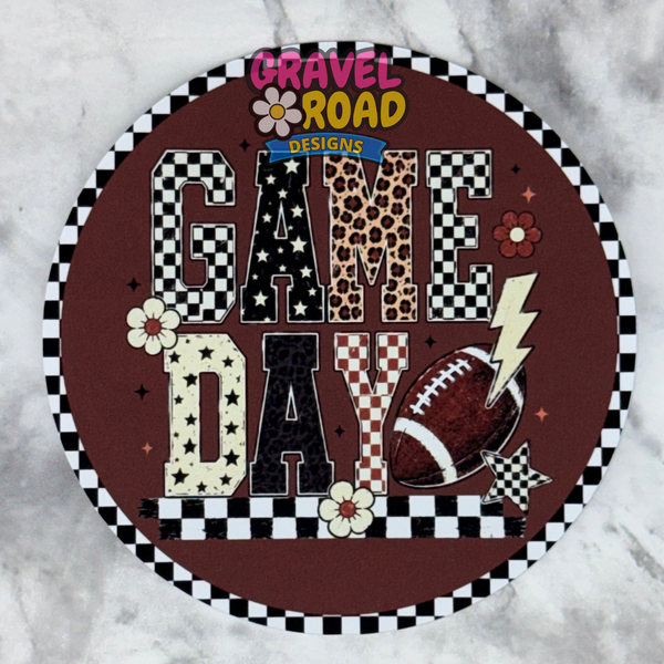 A round cardstock design with a brown background and the words “GAME DAY” in mixed patterned letters, surrounded by a black-and-white checkered border. The sign includes football, lightning bolt, flower, and leopard-print details, and is photographed on a light marble-like surface. A Gravel Road Designs logo is visible at the top.