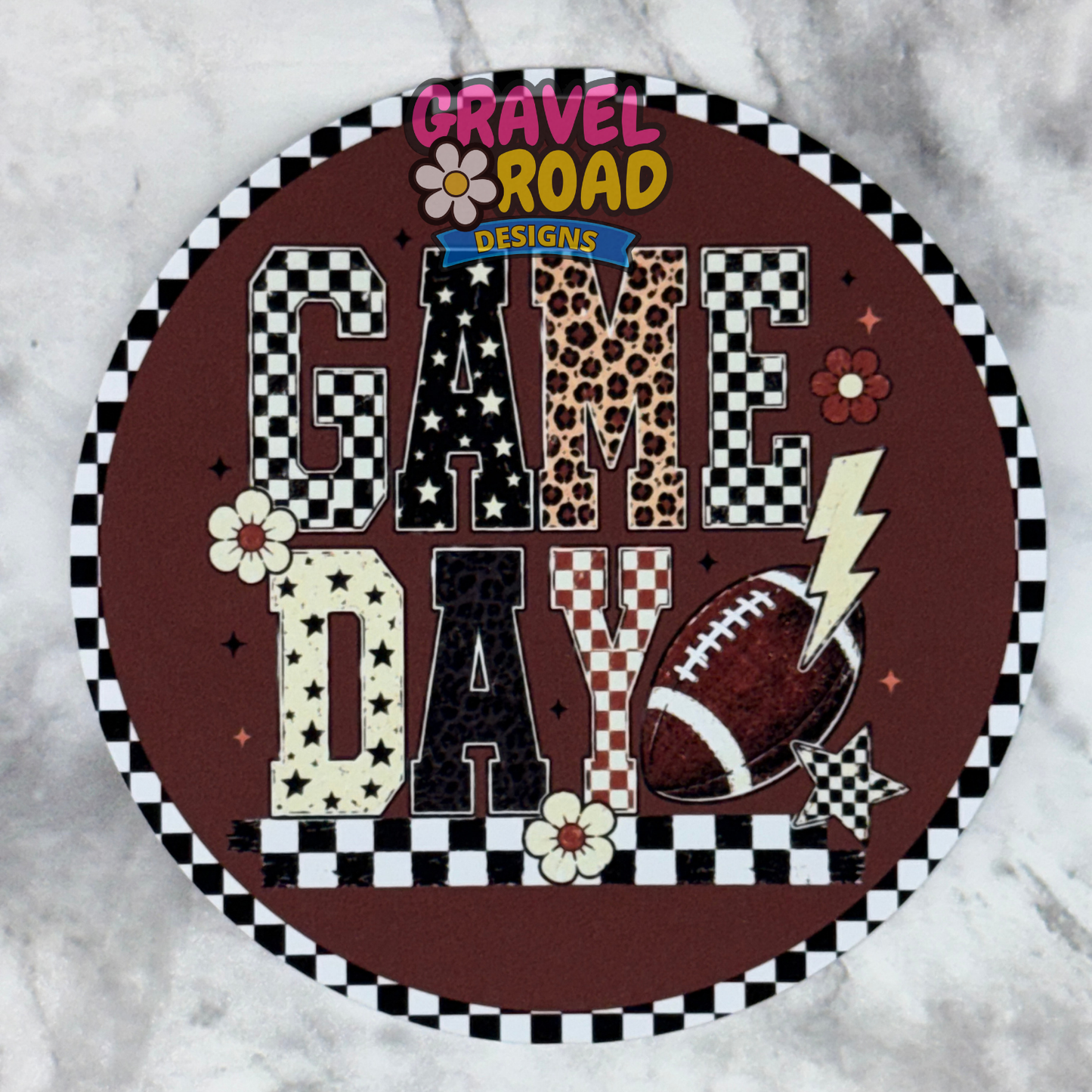 A round cardstock design with a brown background and the words “GAME DAY” in mixed patterned letters, surrounded by a black-and-white checkered border. The sign includes football, lightning bolt, flower, and leopard-print details, and is photographed on a light marble-like surface. A Gravel Road Designs logo is visible at the top.