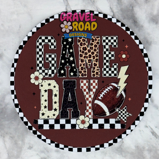 A round cardstock design with a brown background and the words “GAME DAY” in mixed patterned letters, surrounded by a black-and-white checkered border. The sign includes football, lightning bolt, flower, and leopard-print details, and is photographed on a light marble-like surface. A Gravel Road Designs logo is visible at the top.