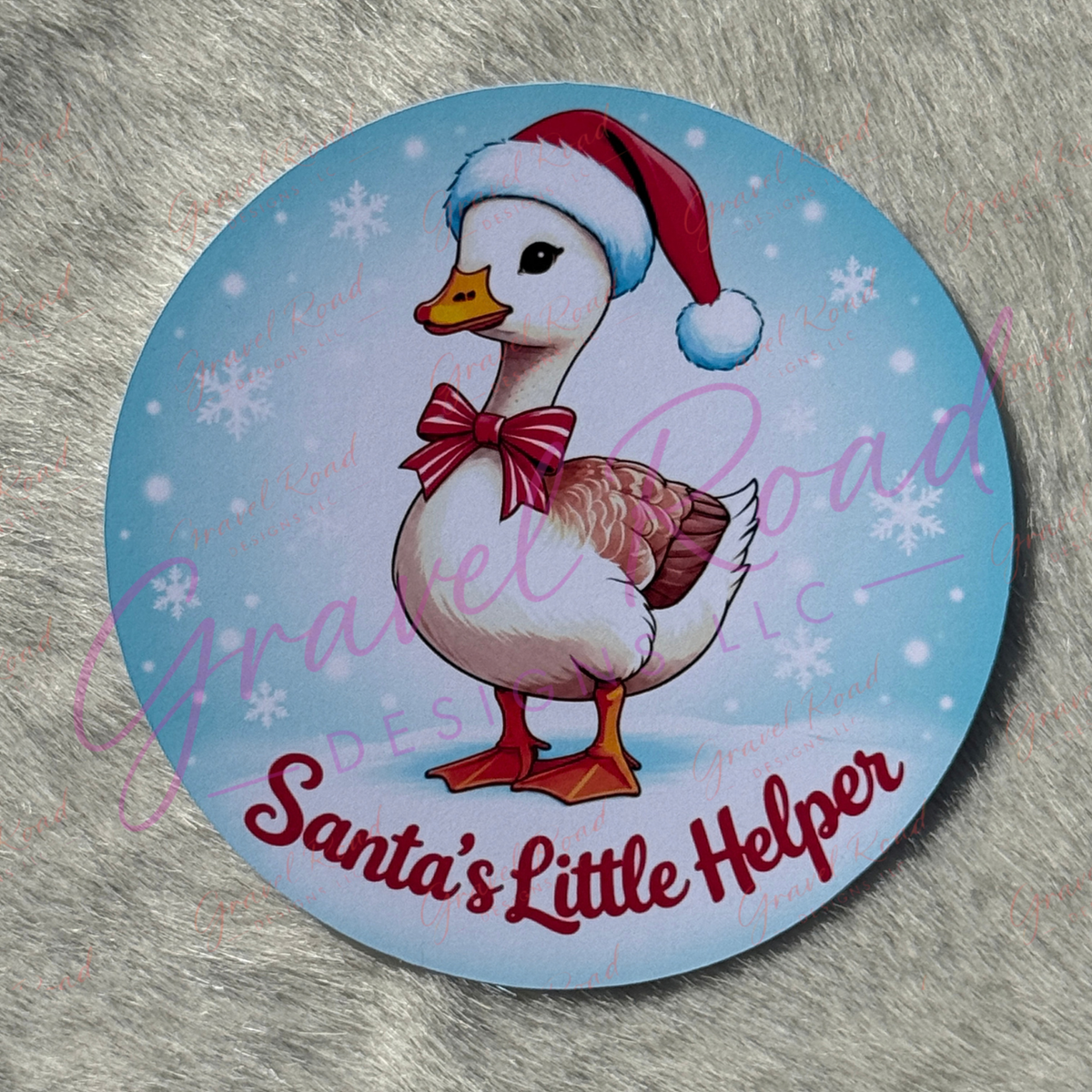 A round printed cardstock featuring an illustrated white duck wearing a red Santa hat and red bow, with the text 'Santa's Little Helper' on a light blue snowy background, photographed on a gray faux-fur surface.