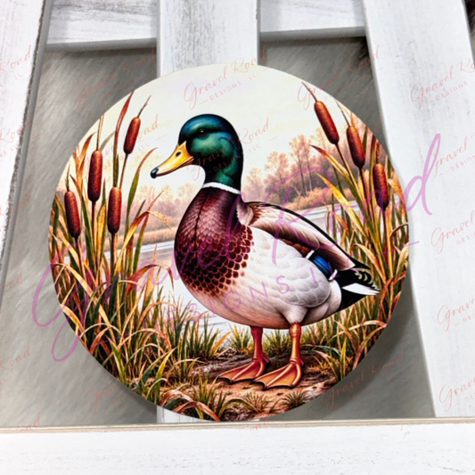 A round cardstock featuring a detailed illustration of a mallard duck standing among cattails, displayed on a white wooden ladder. The plaque has a subtle watermark overlay.