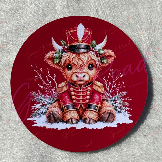 A round cardstock featuring a detailed illustration of a fluffy brown Scottish highland cow dressed in a red military uniform with gold buttons, a red and black hat with a white feather, and a green holly wreath, set against a snowy background.