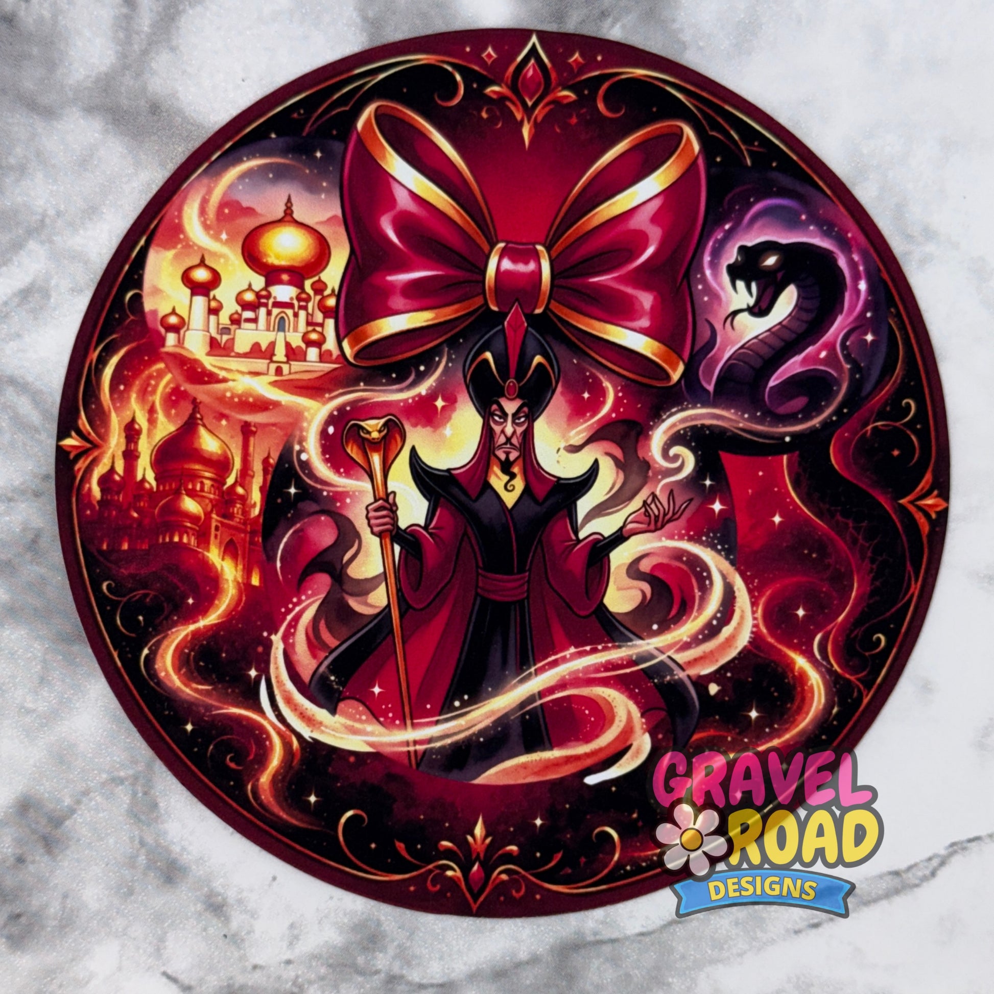A circular cardstock featuring a stylized dark magician or sorcerer surrounded by magical swirls, a large red bow, a serpent, and fantasy castle imagery. The design is presented on a light gray marble-textured background with a 'Gravel Road Designs' logo in the bottom right corner.