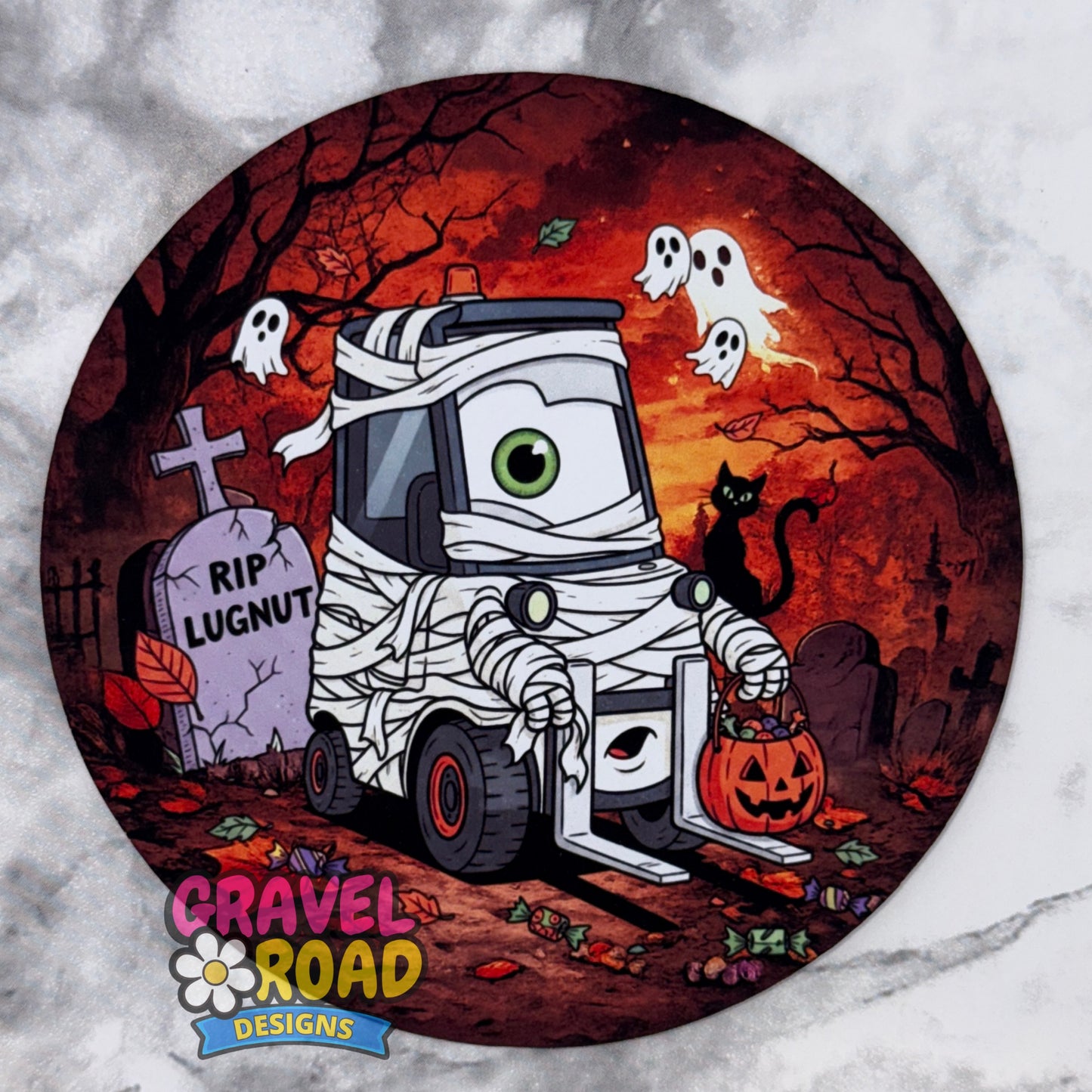A circular Halloween-themed cardstock featuring a cartoon mummy truck with a green eye, a black cat, ghosts, tombstones, and a jack-o'-lantern on an orange autumn background. The coaster is photographed on a white and gray marble surface, with the brand logo 'Gravel Road Designs' printed at the bottom. On a grey marble backdrop.