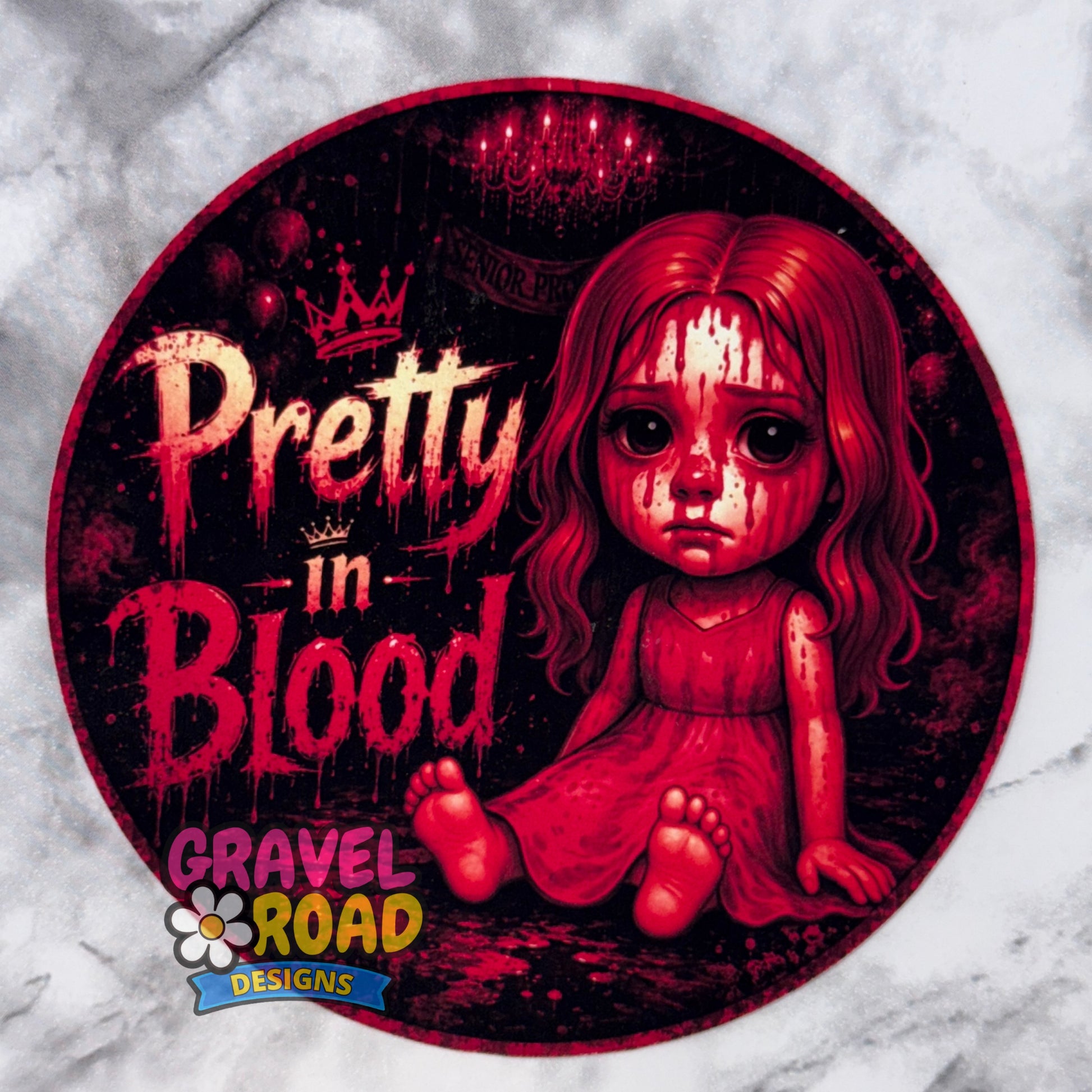 A circular cardstock design featuring a creepy red-haired doll sitting on a dark background with the text 'Pretty in Blood' and a 'Gravel Road Designs' logo at the bottom left, shown on a light marble-patterned background.