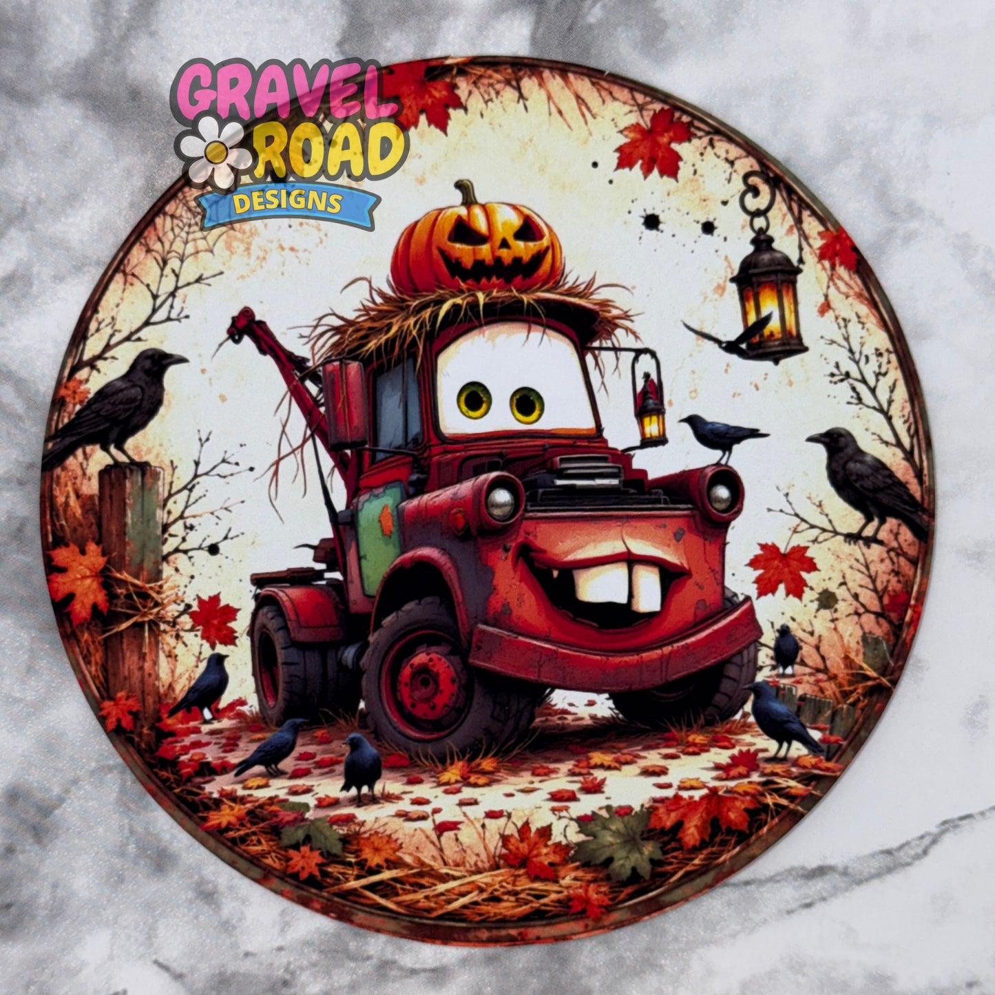 A circular cardstock featuring a cartoon tow truck dressed in autumn Halloween attire, surrounded by crows, leaves, a jack-o'-lantern, and a lantern. The 'Gravel Road Designs' logo is visible in the top left-hand corner. On a grey marble backdrop.