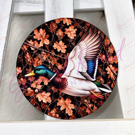 A round cardstock featuring a painted mallard duck in flight over a background of brown and orange autumn leaves, displayed on a white wooden surface.