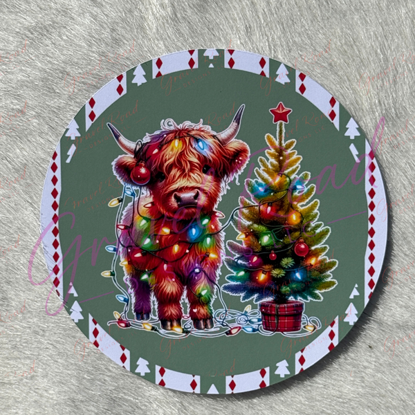 A round Christmas-themed cardstock featuring a shaggy Highland cow and a decorated Christmas tree on a muted green background, with a white border and red diamond accents, shown on a gray textured surface.
