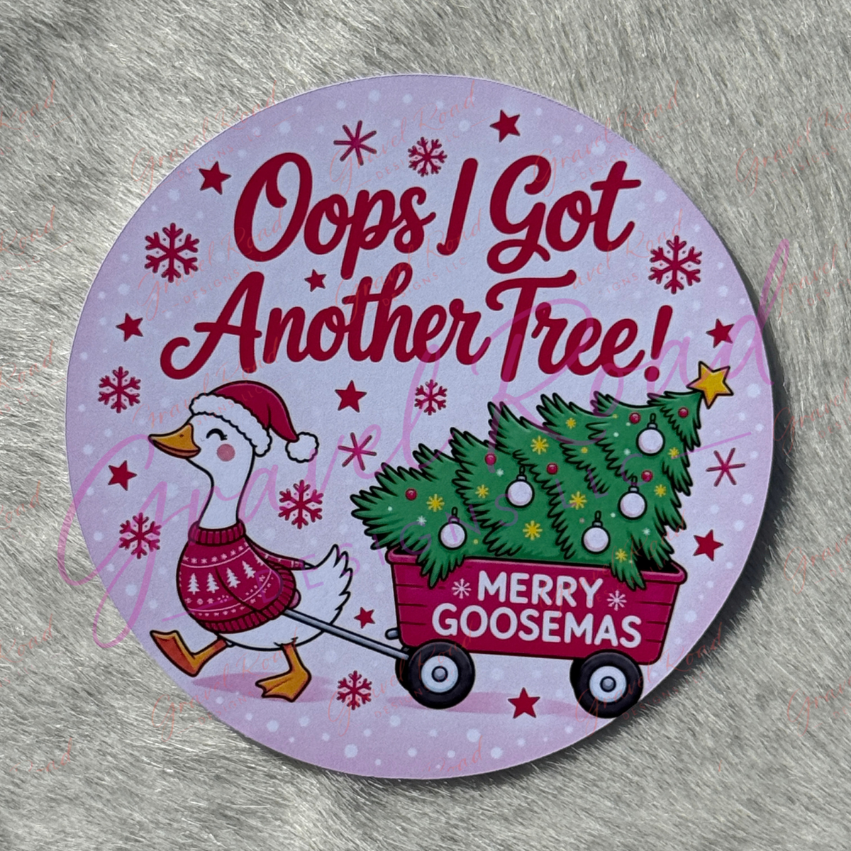 A round printed cardstock featuring a festive illustration of a goose in a red sweater and Santa hat pulling a red wagon loaded with a Christmas tree, with the text 'Oops I Got Another Tree!' and 'Merry Goosemas' on a light lavender background with red stars and snowflakes. Displayed on a faux fur background.